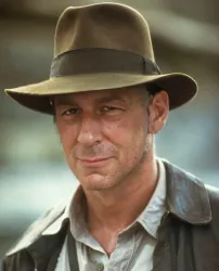 Close-up of a middle-aged man wearing a weathered brown fedora and a rugged leather jacket. He has a confident, subtle smile and his expression portrays a thoughtful demeanor.
