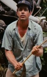 A young man wearing a blue cap and a light blue shirt soiled with dirt stands in front of an airplane wreckage in a tropical forest. He holds a wooden stick, looking upwards with a focused expression.