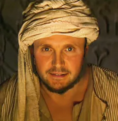 A man dressed in ancient Middle Eastern attire, featuring a white headdress, gazes directly into the camera with a gentle expression. His attire may suggest a cultural representation from biblical times.