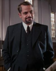 A middle-aged man with a neatly trimmed beard and mustache, wearing a dark formal suit with a white shirt and dark tie, stands in a room with large windows. His expression is thoughtful and slightly amused.